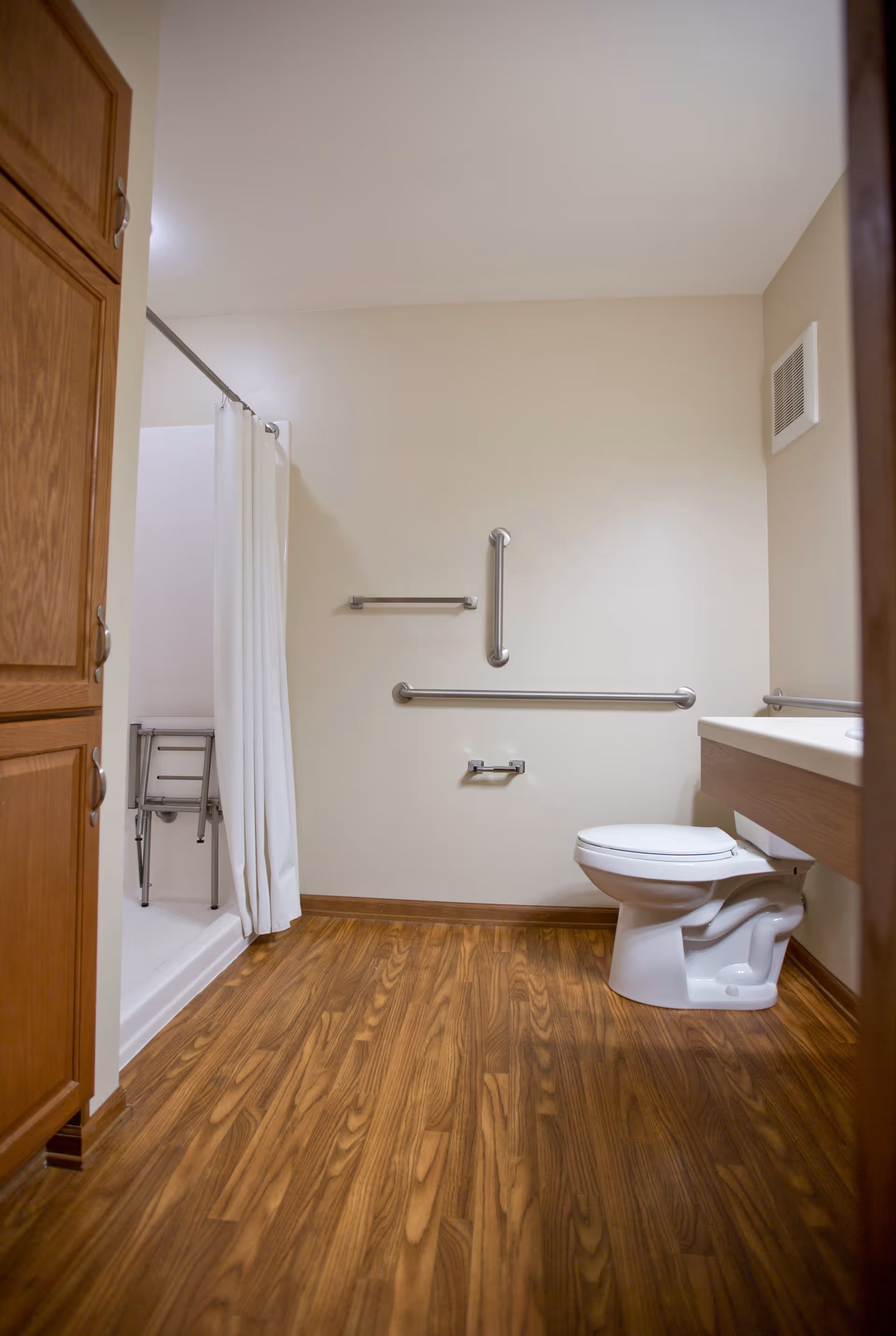 Accessible bathroom with wood-look flooring, a toilet, wall-mounted grab bars, and a shower with a curtain and shower chair.