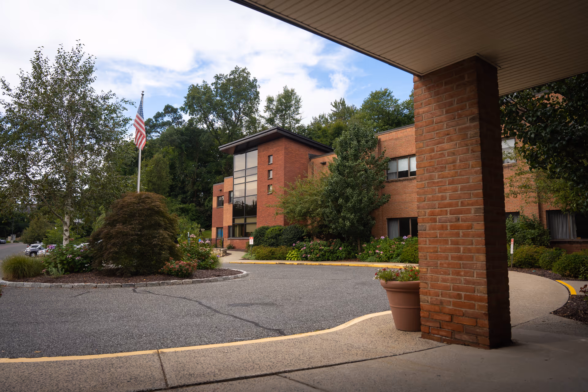 View of the exterior of a brick senior living facility building with large windows, surrounded by trees and landscaping. There is a circular driveway with a flagpole displaying the American flag in the center. The photo is taken from under a covered entrance with a brick pillar and a large flower pot.