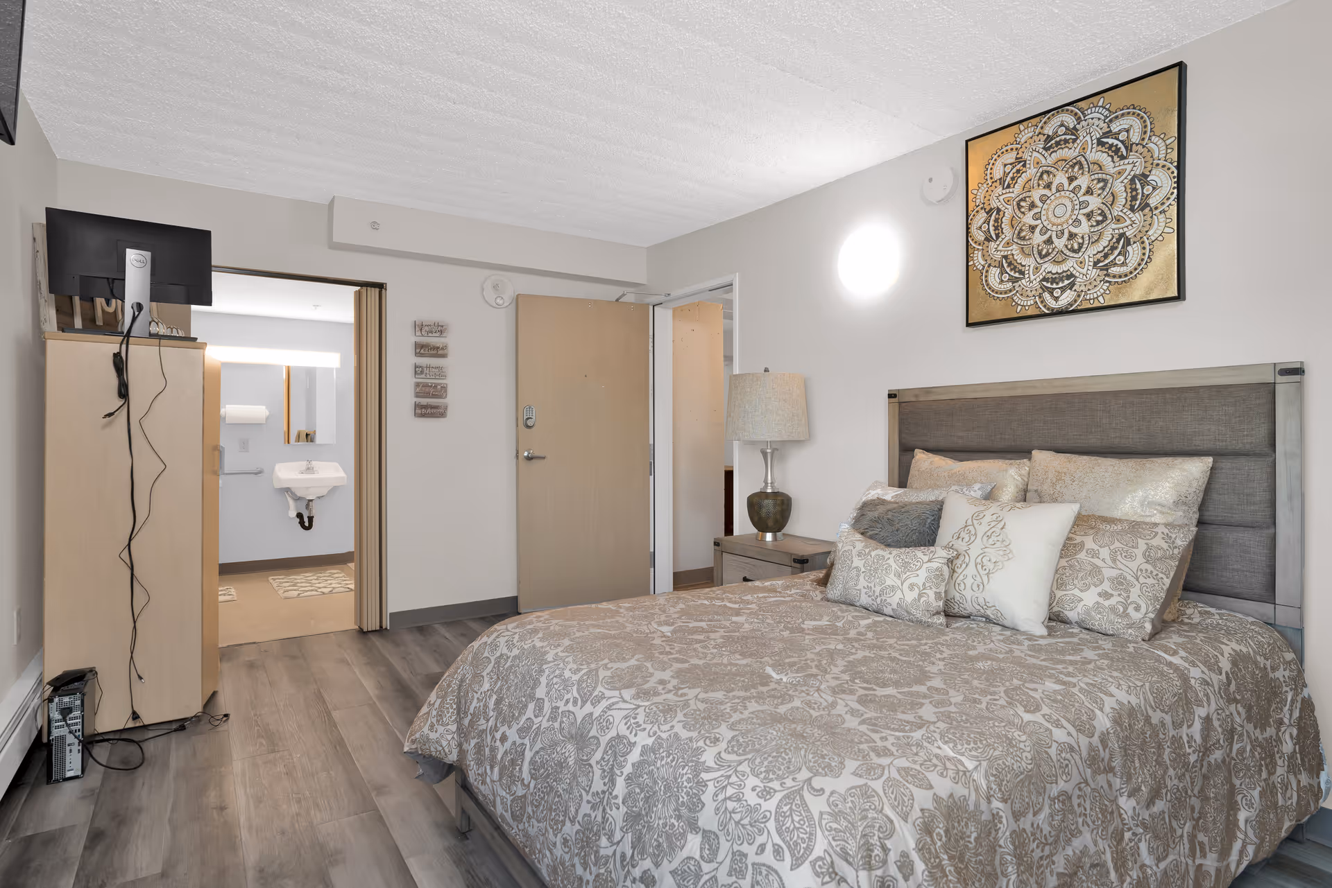 A bedroom in an assisted living facility featuring a bed with patterned bedding and multiple pillows, a nightstand with a lamp, a wall-mounted decorative artwork above the bed, a wooden dresser with a TV on top, and an open door leading to a bathroom with a sink and mirror visible.