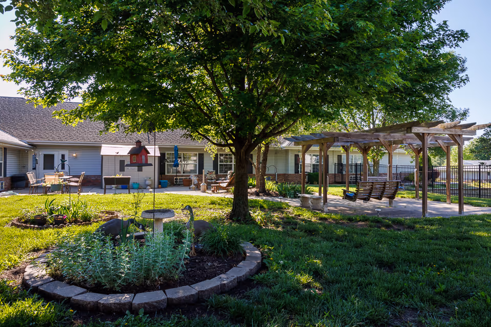 A sunny outdoor garden area at Homestead Assisted Living of Auburn featuring a large tree in the center, a circular flower bed with plants and garden decorations, a bird feeder, and a wooden pergola with two wooden swings. The background shows a single-story building with a patio area furnished with chairs and tables.