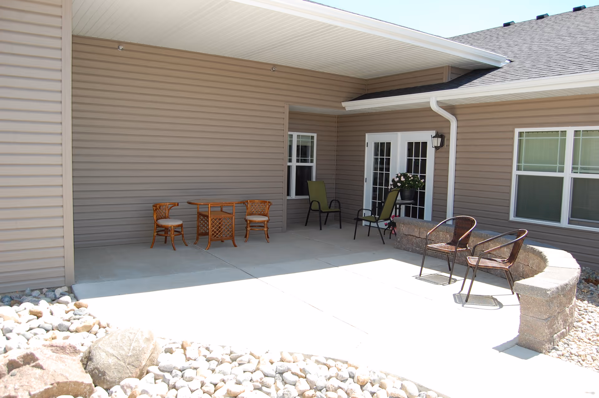 Outdoor patio area with beige siding walls, a concrete floor, and a curved stone bench. There are several chairs including two green cushioned chairs, two wicker chairs with cushions, and two metal chairs. A small wooden table is placed between the wicker chairs, and a potted plant is near the double glass doors.