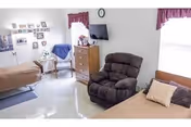 Bright senior bedroom with two beds, a recliner, a dresser with a wall-mounted TV, and personal photos on the wall.