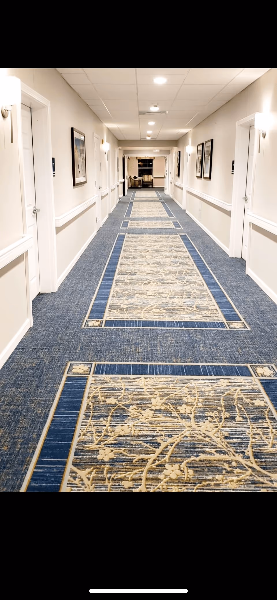 A long, well-lit hallway in an assisted living facility with patterned blue and beige carpet runners. The walls are light-colored with white trim and handrails. Several closed white doors line both sides of the hallway, and framed pictures hang on the walls. At the end of the hallway, there is a seating area with chairs and a window.
