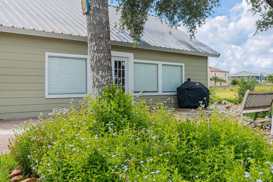 Exterior view of a single-story building with large windows, a tree, flowering shrubs, a covered grill, and a bench.