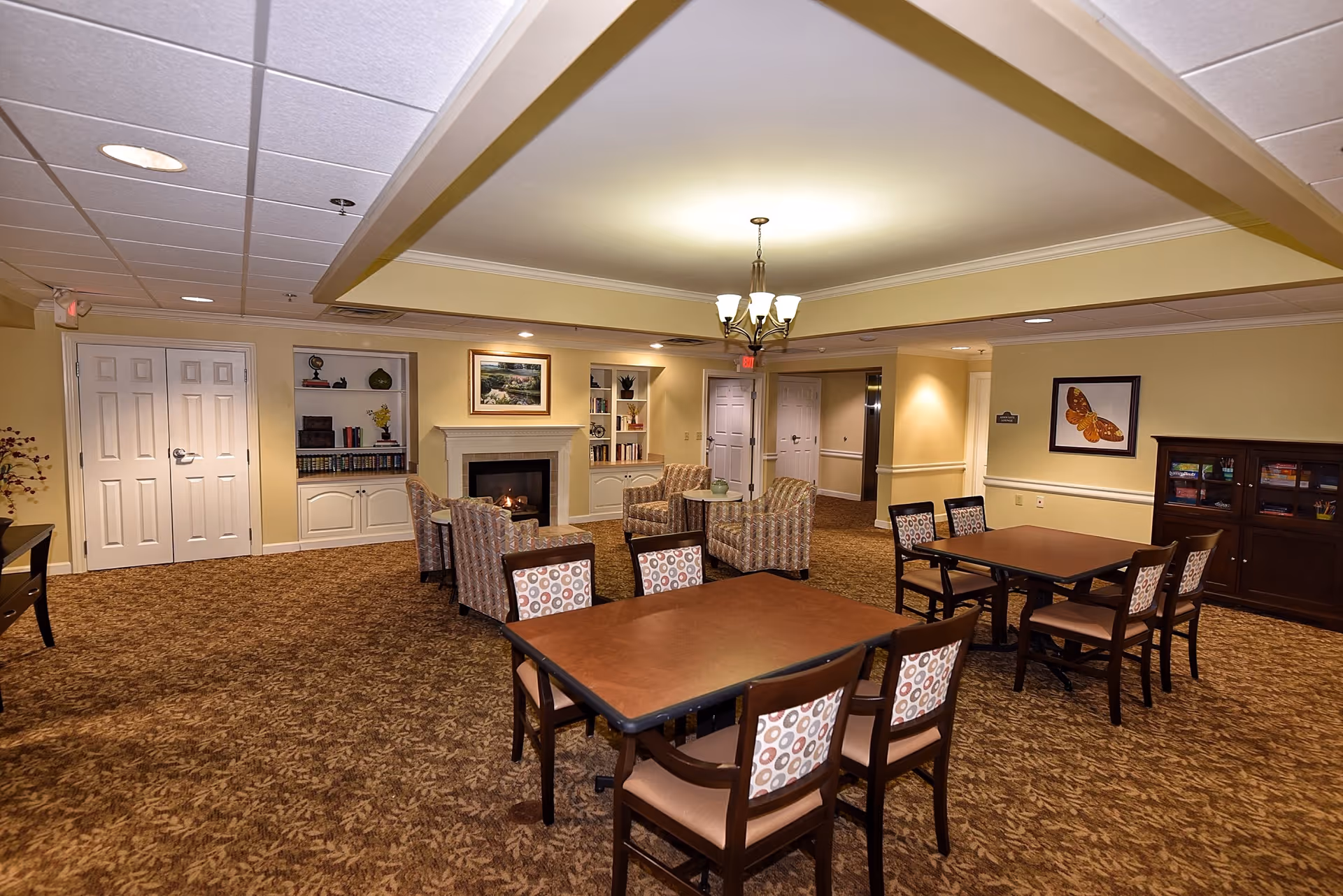 A cozy senior living common area with two tables and chairs in the foreground, a seating area with four armchairs around a small table near a fireplace, built-in shelves with books and decor, warm yellow walls, carpeted floor, and a chandelier hanging from the ceiling.