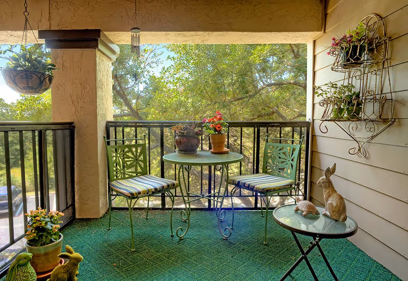 A cozy outdoor balcony with a small green metal bistro table, two cushioned chairs, potted plants, and decorative animal statues overlooking trees.