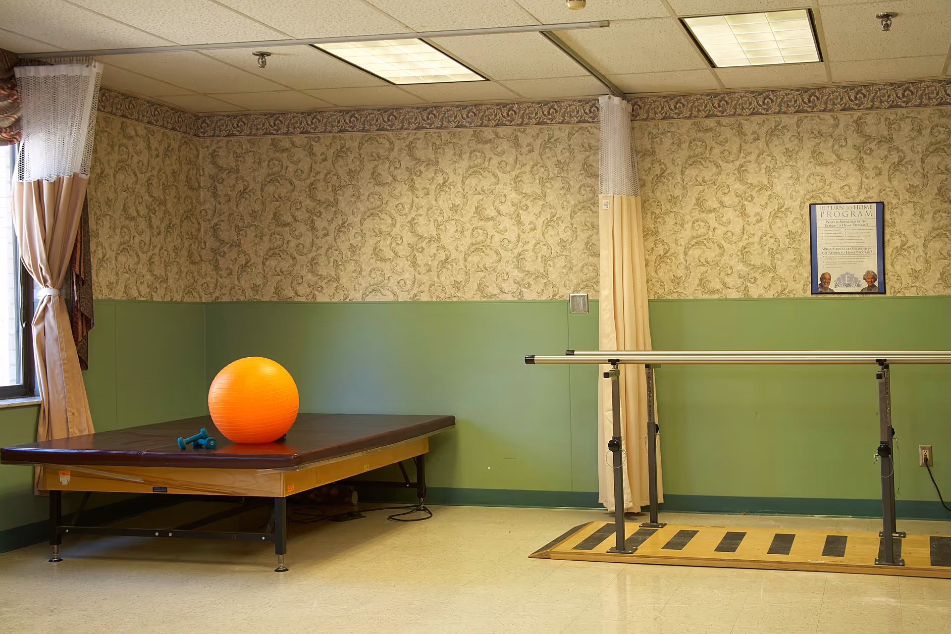 A therapy or exercise room with a padded table holding an orange exercise ball and two small blue dumbbells. There are parallel bars for walking exercises on the right side, a window with curtains on the left, and a framed informational poster on the wall. The walls are green on the lower half and patterned wallpaper on the upper half.