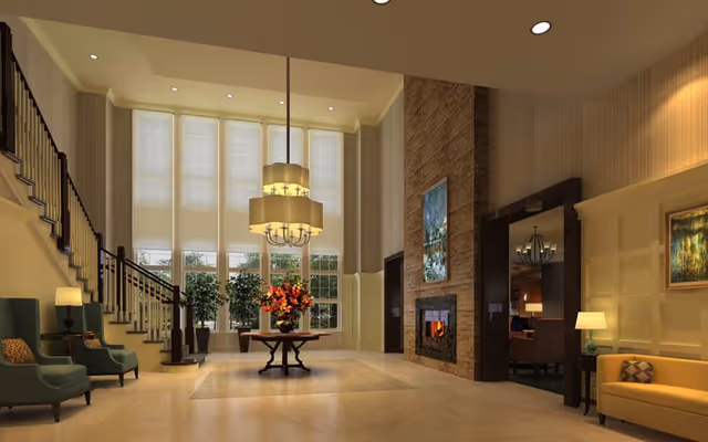 Spacious and elegant living room area with high ceilings and large windows covered with white blinds. The room features a central round table with a floral arrangement, a modern chandelier hanging above it, a stone fireplace with a painting above, green armchairs on the left near a staircase, and a yellow sofa with a lamp and painting on the right.