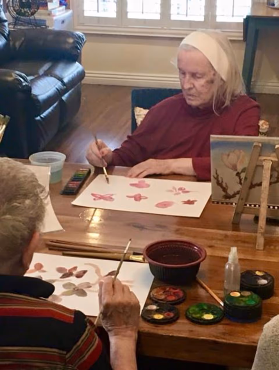 Two elderly individuals seated at a wooden table engaged in watercolor painting. One person is painting pink butterflies and flowers on a sheet of paper, while the other is painting similar floral designs. Various paint palettes, brushes, and a water container are on the table. The setting appears to be a cozy indoor room with a leather chair and a window with white shutters in the background.