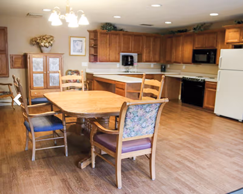 A spacious kitchen and dining area with wooden cabinets, a white refrigerator, a black microwave, and a black dishwasher. There is a wooden dining table with four chairs, one of which has floral upholstery. The floor is wooden, and there is a cabinet with glass doors and a flower arrangement on top against the wall.