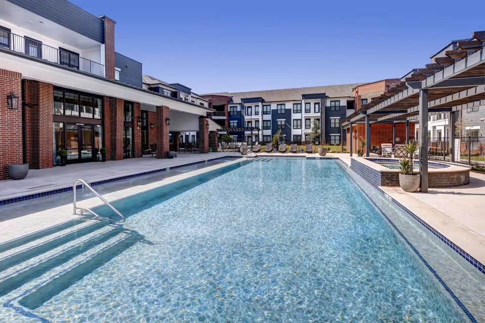 Outdoor swimming pool and deck area with lounge chairs in front of a multi-story brick and siding senior living building.