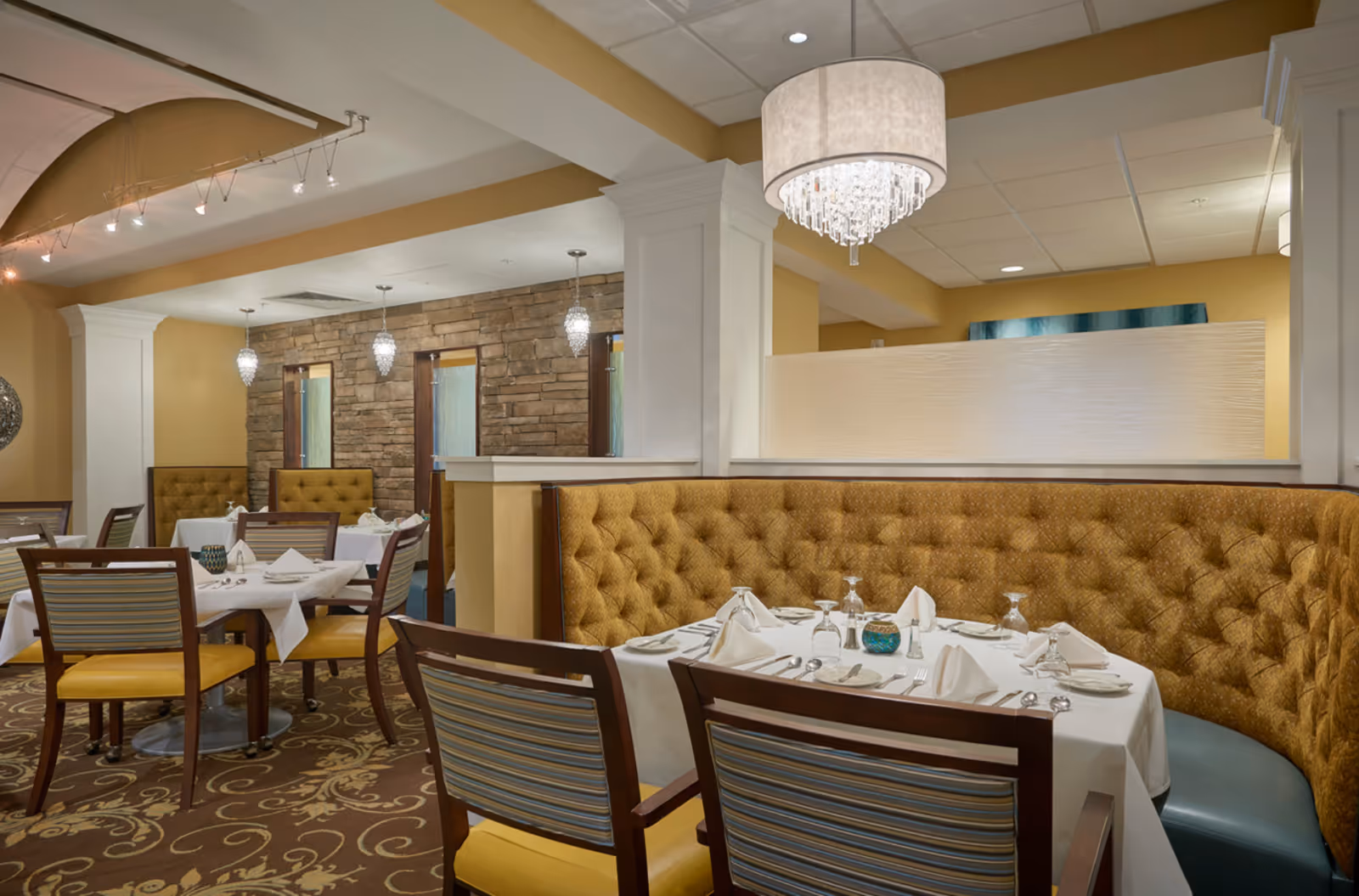 Elegant dining area with tables set with white tablecloths, napkins, plates, and glassware. The seating includes mustard-colored tufted banquettes and wooden chairs with striped cushions. The room features warm yellow walls, a decorative carpet, stone accent wall, and modern pendant and chandelier lighting.