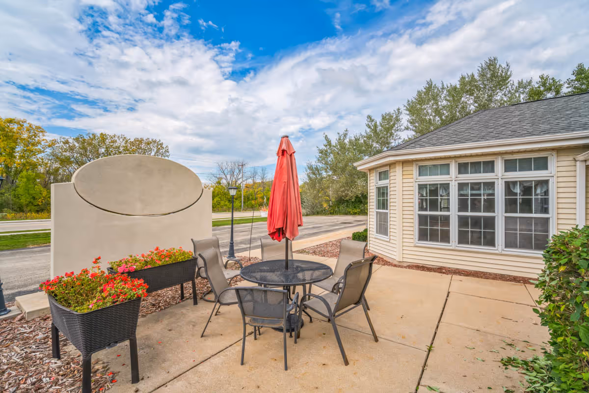 Outdoor patio area with a round metal table and four chairs, a closed red umbrella in the center, two planters with red flowers, a beige building with large windows, and a blank beige sign. Trees and a partly cloudy sky are visible in the background.