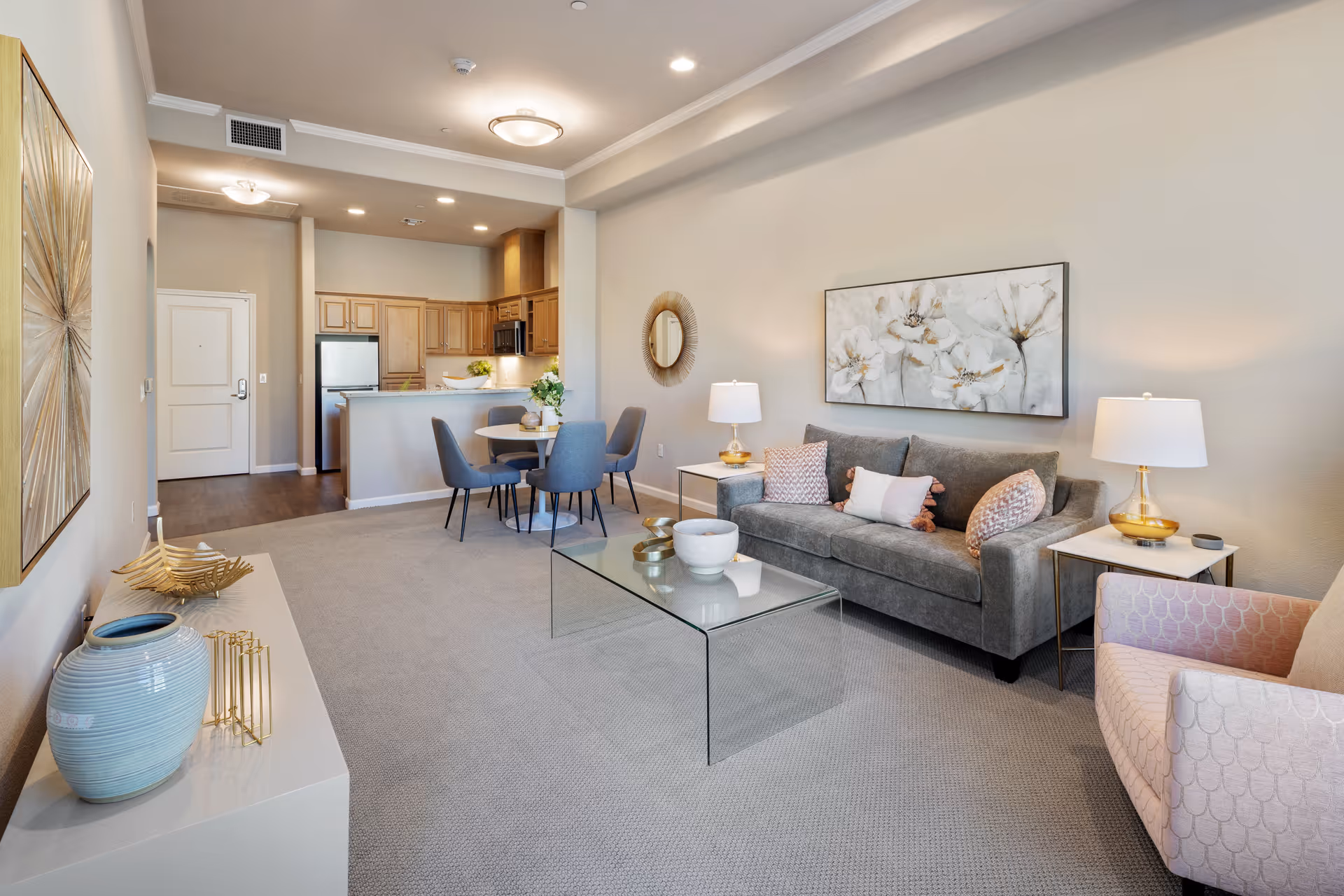 A bright and modern living room and kitchen area in a senior living facility. The living room features a gray sofa with decorative pillows, a glass coffee table, two side tables with lamps, and a pink armchair. A large floral painting hangs on the wall above the sofa. The kitchen area includes wooden cabinets, a refrigerator, microwave, and a small round dining table with four gray chairs. The room has beige walls, carpeted flooring in the living area, and wood flooring in the kitchen area.