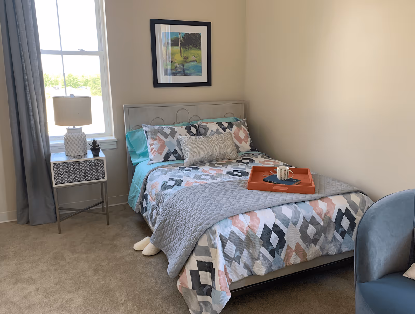 Neatly made single bed with geometric bedding, a nightstand and lamp beside a window in a small bedroom.