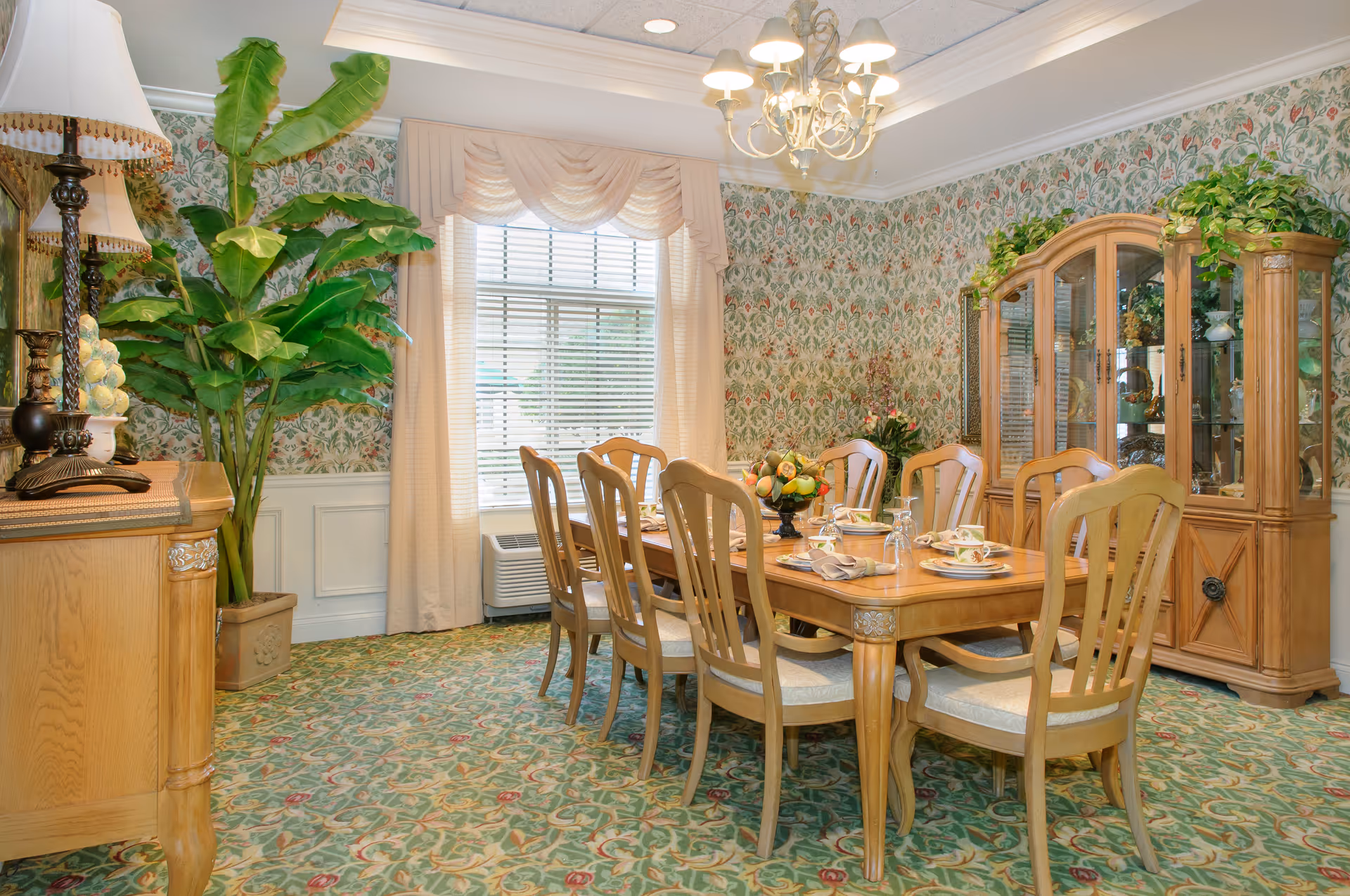 A traditional dining room with a wooden dining table set for six, surrounded by matching wooden chairs. The room features floral wallpaper, a large window with cream-colored curtains, a chandelier overhead, a wooden china cabinet with glass doors displaying decorative items, and a large potted plant in the corner. The carpet has a floral pattern that complements the wallpaper.