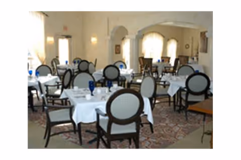 A dining room with multiple tables covered in white tablecloths, each set with blue water glasses and white cups. The room has upholstered chairs with dark wooden frames, large windows with sheer curtains, and soft lighting on the walls.