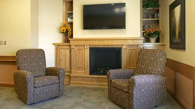 A cozy living room area with two patterned armchairs facing a wooden fireplace mantel. Above the fireplace is a mounted flat-screen TV. Built-in shelves on either side of the fireplace hold books and decorative plants. The walls are painted a light yellow, and there is a framed picture on the right wall.