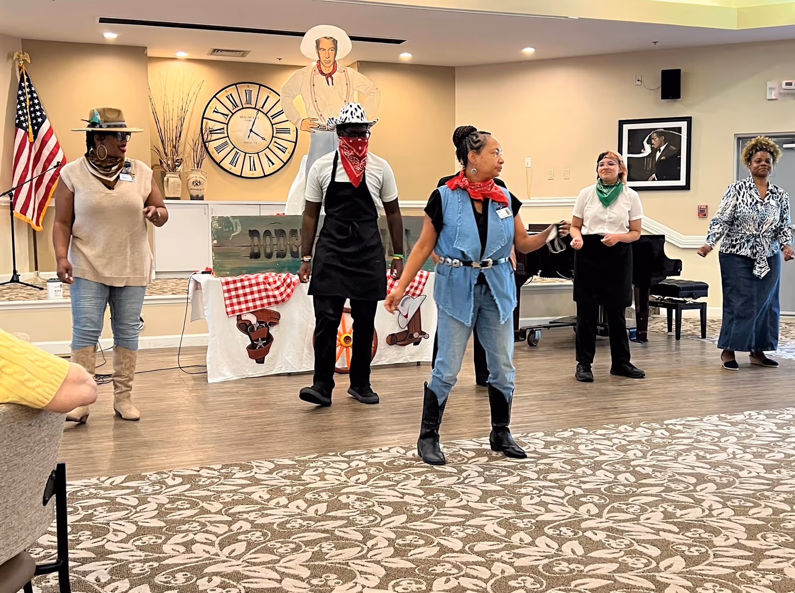 A group of five adults dressed in western-themed attire, including hats, bandanas, and boots, standing and interacting in a spacious room with patterned carpet and beige walls. Behind them is a large clock, an American flag, and a cutout figure of a cowboy. The room appears to be a community or activity space.