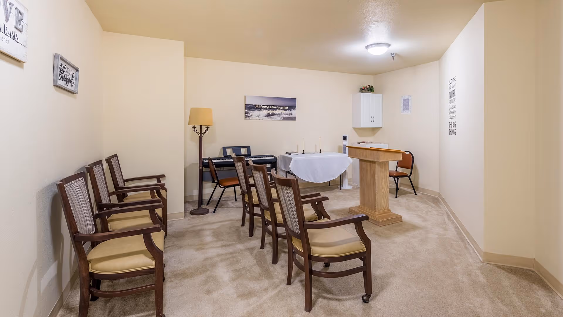 A small room with beige walls and carpeted floor set up for a gathering or service. There are two rows of wooden chairs with cushioned seats facing a wooden podium and a table covered with a white cloth. A keyboard and a floor lamp are positioned against the back wall. Wall decorations include framed inspirational quotes and a small cabinet mounted on the wall.