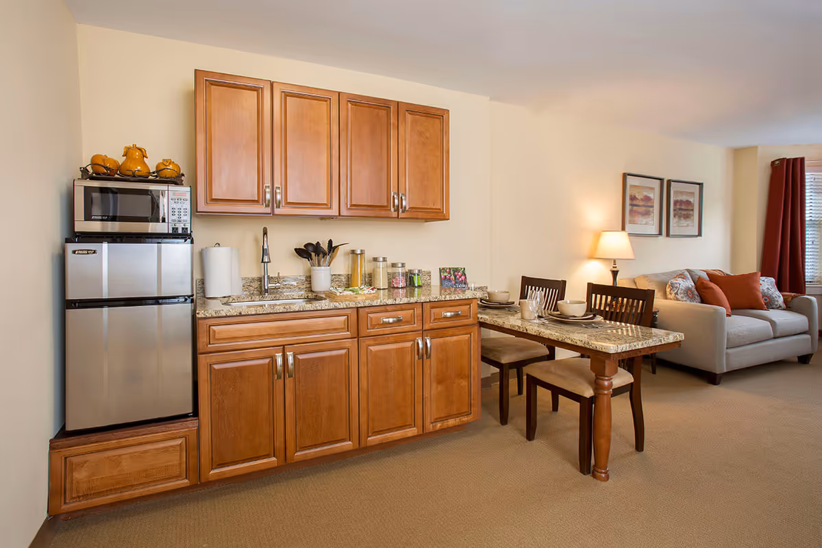 A cozy senior living kitchenette and dining area featuring wooden cabinets, a small stainless steel refrigerator with a microwave on top, a granite countertop with a sink, kitchen utensils, and jars. Adjacent to the kitchenette is a granite dining table set with two chairs, bowls, and plates. In the background, there is a living room area with a beige sofa, decorative pillows, a table lamp, framed artwork on the wall, and a window with red curtains.