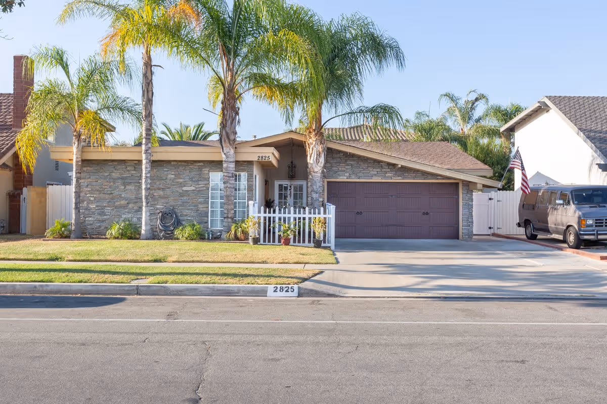 Front exterior view of a single-story house with stone facade, a brown garage door, three tall palm trees in the front yard, a small white picket fence with potted plants, and a driveway with a parked van displaying an American flag.