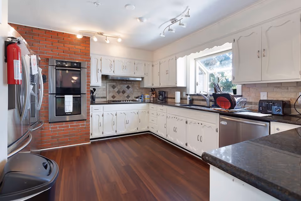 A bright kitchen with white cabinets, dark countertops, and wooden flooring. The kitchen features a double oven built into a brick wall, a stainless steel refrigerator, a dishwasher, a sink under a window, and various kitchen appliances on the counters.