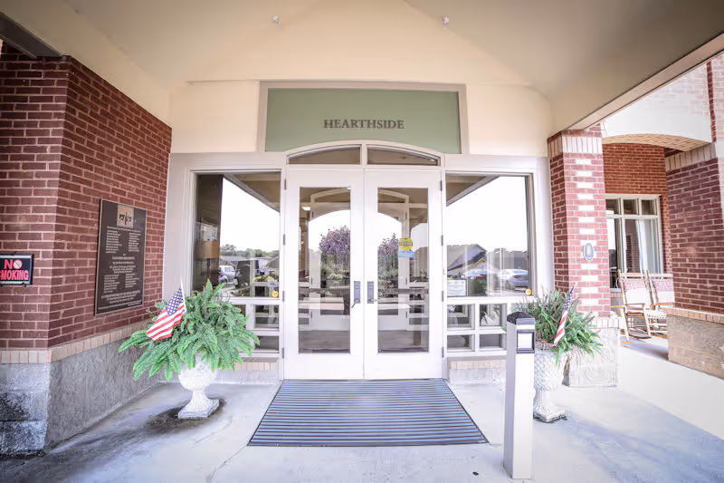 Front entrance of Hearthside Senior Living with double glass doors under a sign reading "HEARTHSIDE", brick columns, and potted plants displaying American flags.