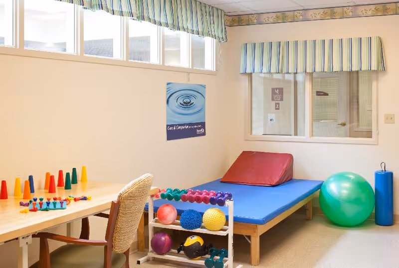 Activity/therapy room with a padded mat and wedge, exercise balls, hand weights, and a table with colorful cones and toys.