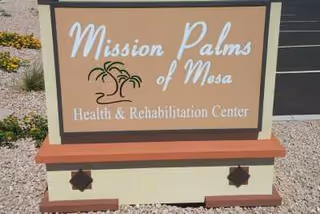 A sign for Mission Palms of Mesa Health & Rehabilitation Center placed outdoors on a gravel surface with some plants visible in the background.