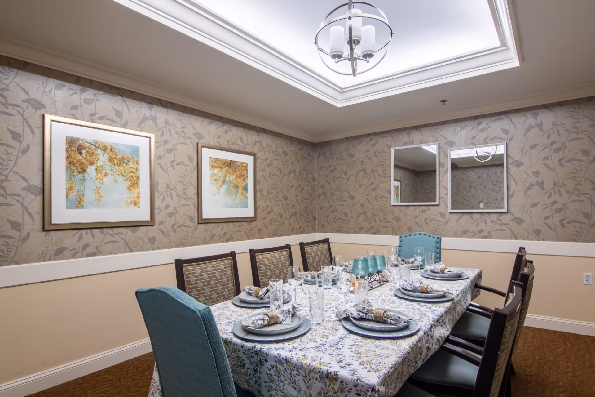 A dining room with a rectangular table covered with a floral tablecloth set for eight people. The table has plates, napkins with rings, glasses, and teal-colored goblets. The room has patterned wallpaper with a floral design, two framed pictures of flowers on one wall, and two mirrors on another wall. There is a ceiling light fixture and a skylight above the table.