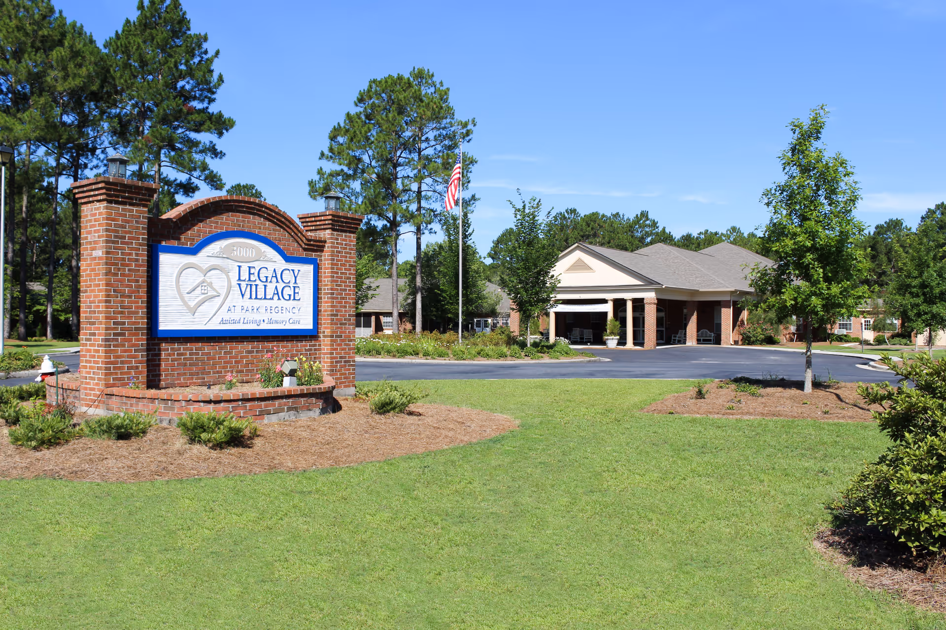 Exterior view of Legacy Village at Park Regency senior living facility with a large brick sign in the foreground and a building with a covered entrance in the background, surrounded by green grass, trees, and a clear blue sky.