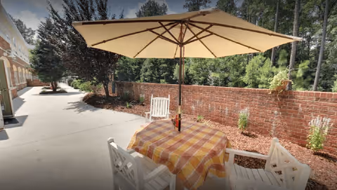 Outdoor patio with a table covered by a checkered cloth under a large umbrella and white chairs beside a brick wall.