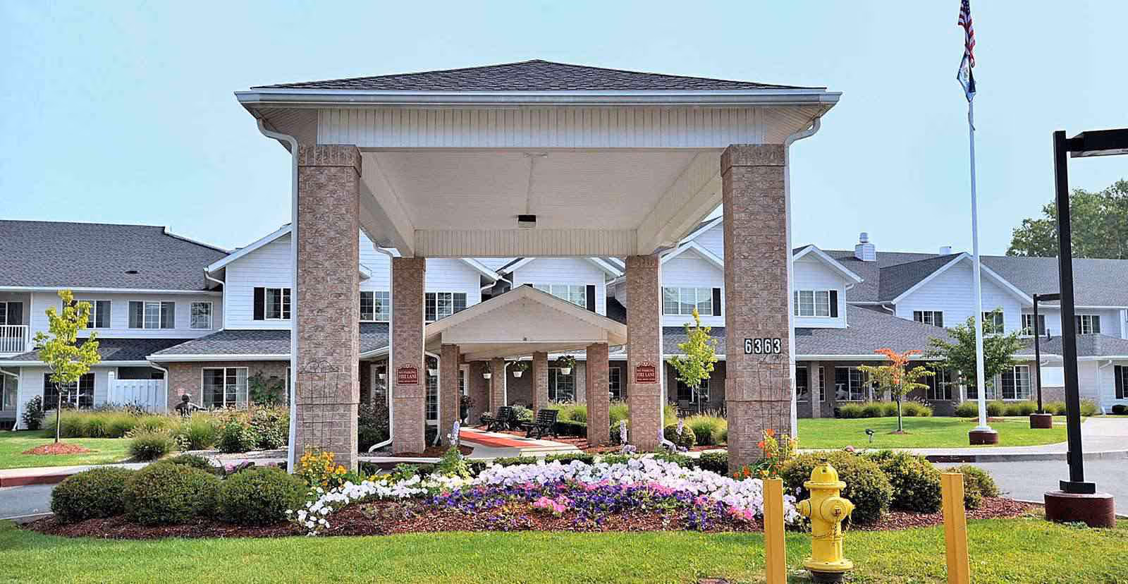 Front entrance of a two-story senior living facility with a covered porte-cochere, landscaped flower beds, and a yellow fire hydrant.