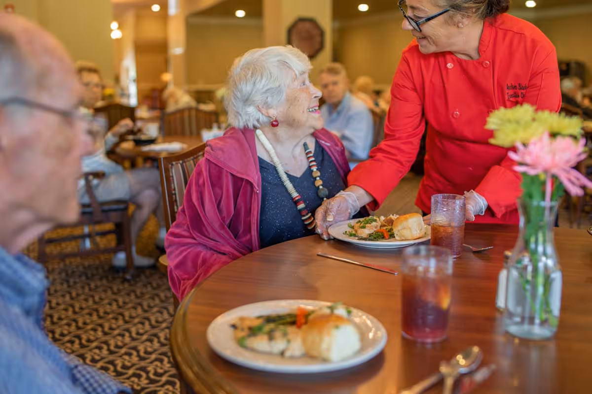 An elderly woman sitting at a dining table in a communal dining room, smiling and looking up at a staff member in a red uniform who is serving her a plate of food. Other elderly people are seated at tables in the background, and there are glasses of iced tea and a vase with flowers on the table.