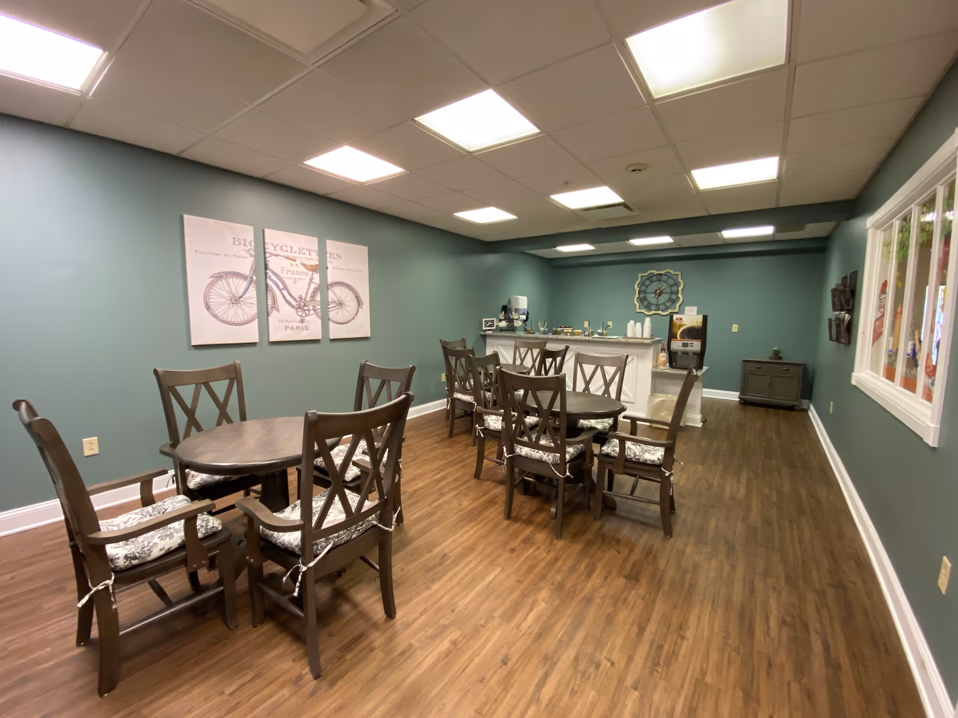 A dining room with two wooden tables, each surrounded by six cushioned chairs. The walls are painted green, and there is a three-panel artwork of a bicycle on one wall. At the far end of the room, there is a counter with a coffee machine, cups, and other beverage supplies. The floor is wooden, and the ceiling has recessed lighting panels.