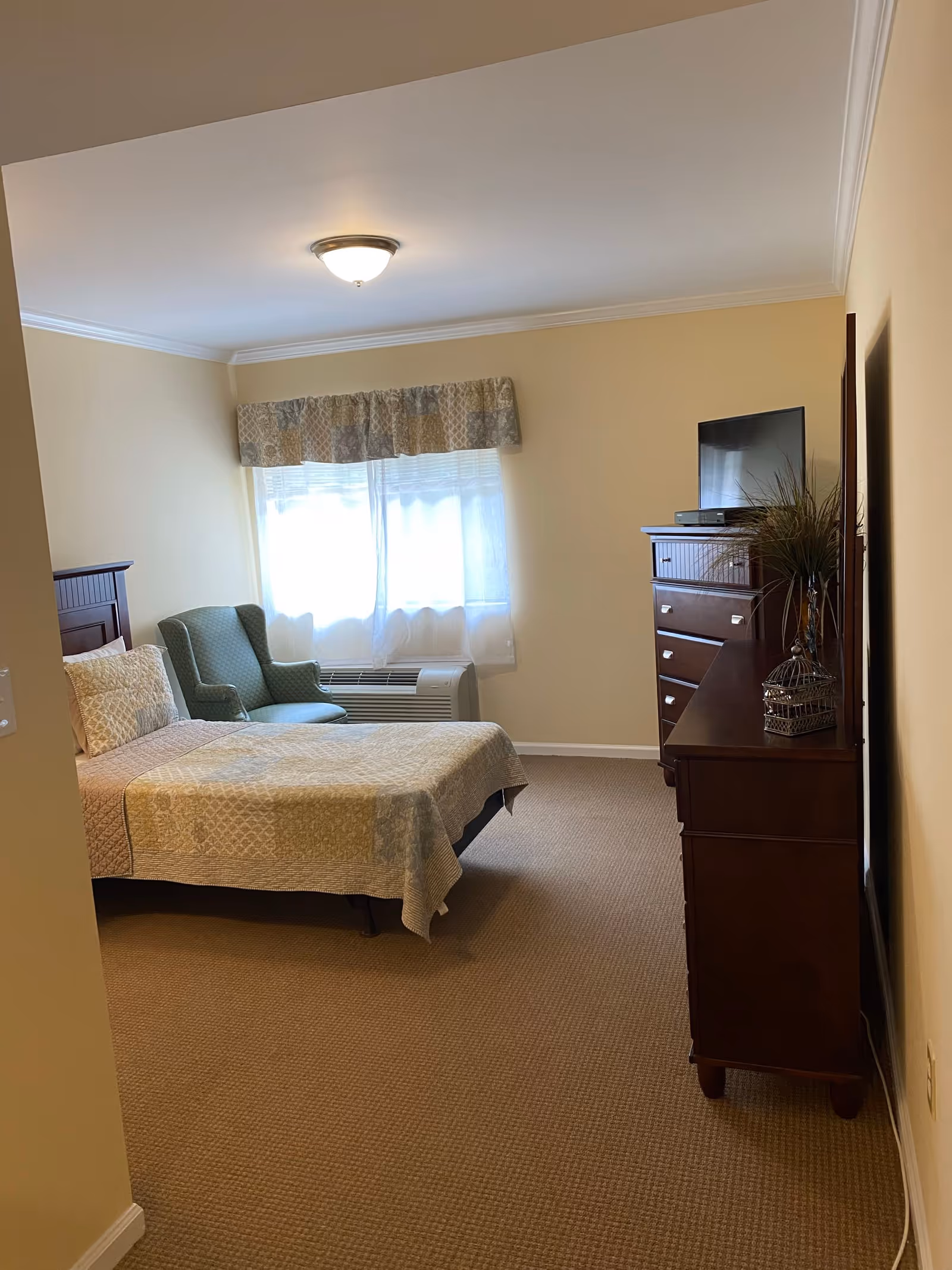 Small furnished bedroom with a bed, an upholstered armchair by a window, and a dresser topped by a TV.