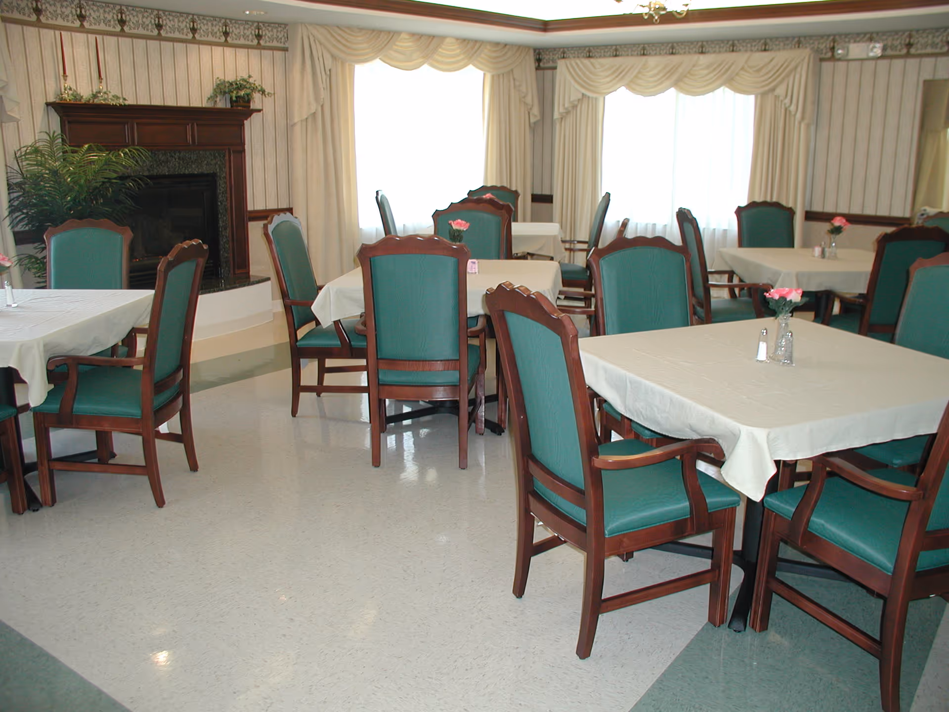 Dining room with multiple tables covered in white tablecloths, green-upholstered chairs, small flower vases, large draped windows and a fireplace.