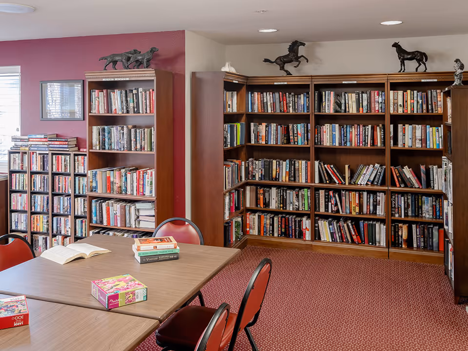 A cozy community library room with wooden bookshelves filled with books, tables and red chairs, and books and puzzles on the tables.