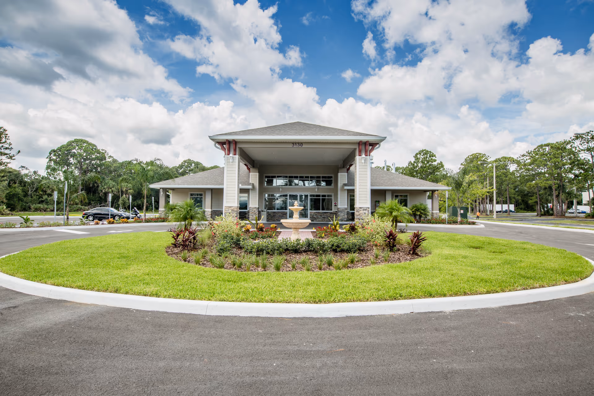 Front exterior view of a single-story building with a covered entrance, surrounded by a circular driveway and landscaped greenery with a small fountain in the center. The sky is partly cloudy.