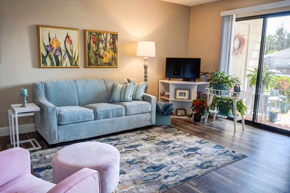 A cozy living room with a light blue sofa adorned with patterned pillows, a pink armchair and matching ottoman, a floor lamp, and a small white side table with a candle. Two framed floral paintings hang on the wall above the sofa. A TV sits on a white corner shelf unit decorated with framed photos and plants. Large sliding glass doors open to an outdoor patio with greenery visible outside.
