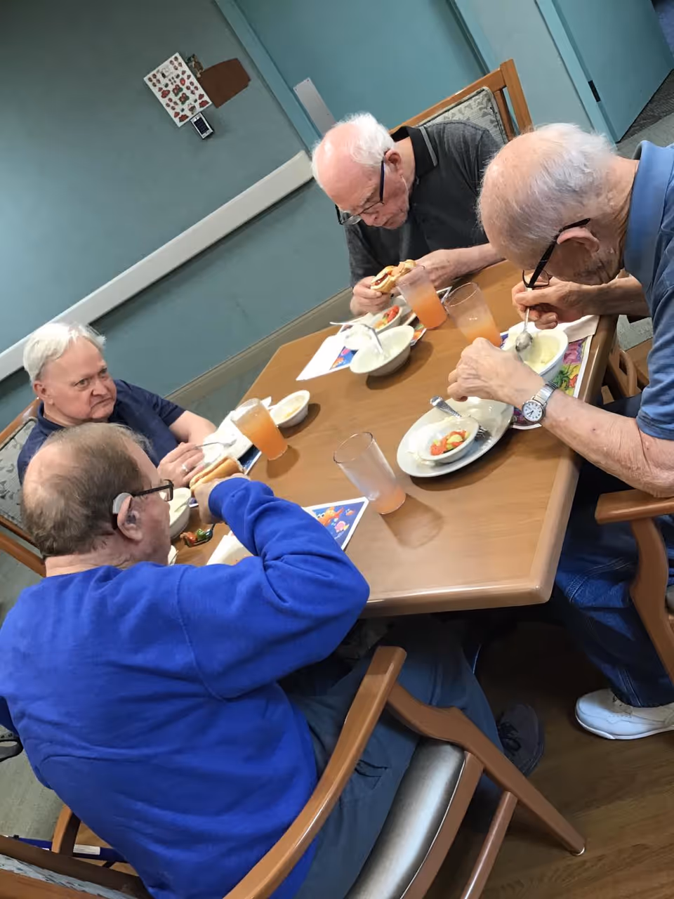 Four elderly men sitting around a wooden table in a dining area, eating a meal that includes soup and sandwiches, with glasses of orange-colored drink on the table. The room has light blue walls and simple decor.