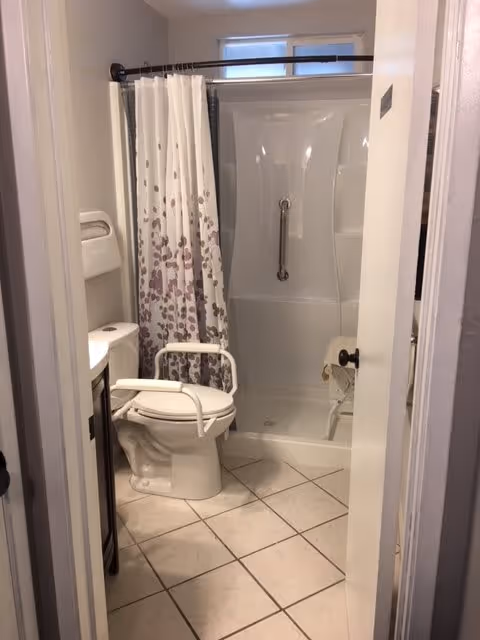 A small bathroom with a white toilet equipped with safety rails, a shower with a white curtain featuring a purple leaf pattern, a grab bar inside the shower, and tiled floor. The bathroom door is partially open.