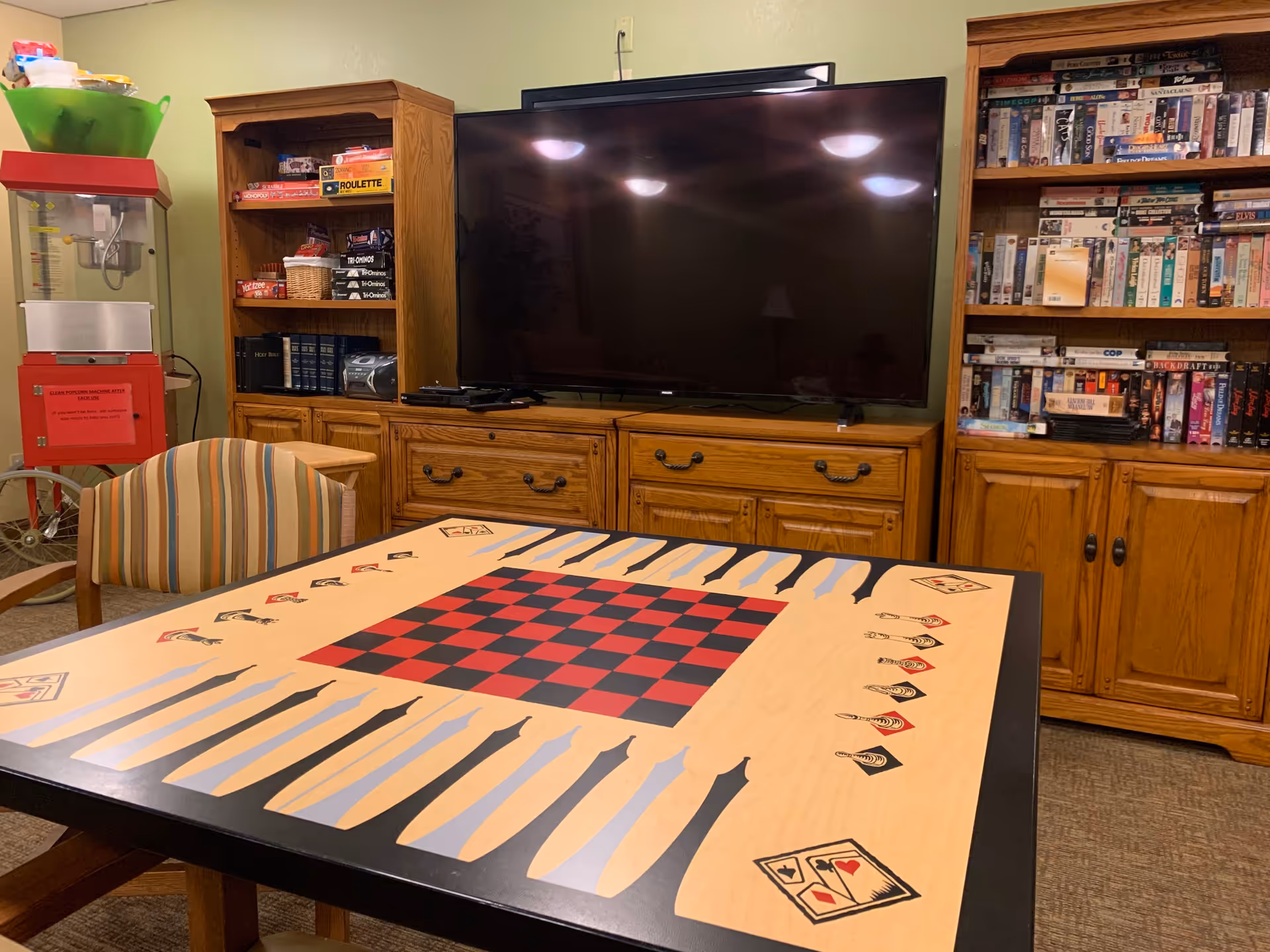 A cozy game room with a large TV mounted on a wooden cabinet, shelves filled with board games and VHS tapes, a popcorn machine on the left, and a table with a checkerboard and backgammon design in the foreground. A striped chair is positioned next to the table.