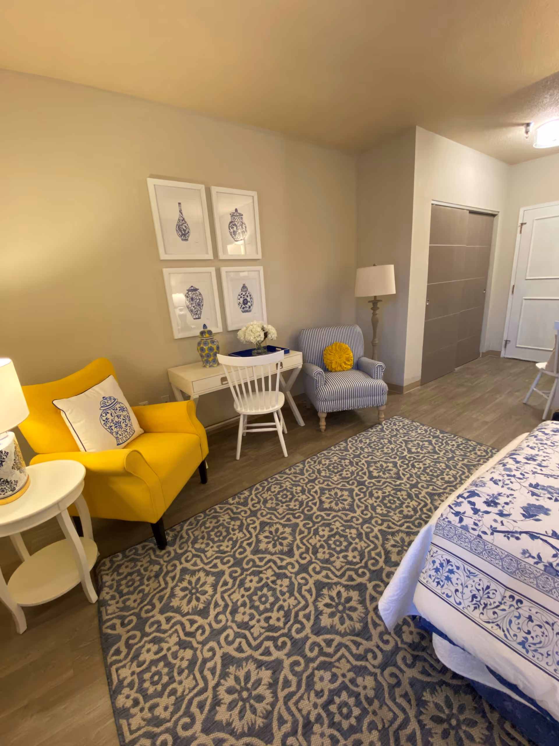 A cozy bedroom corner featuring a yellow armchair with a decorative pillow, a white side table with a lamp, a small white desk with a white chair, and a blue and white striped armchair with a yellow round pillow. The wall above the desk has four framed blue and white vase prints. The floor has a patterned blue and beige rug, and part of a bed with blue and white floral bedding is visible.