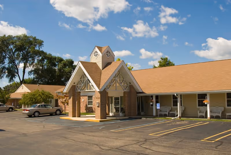 Exterior view of a single-story building with a peaked entrance and brick pillars, surrounded by a parking lot with a few parked cars and some trees in the background under a partly cloudy sky.