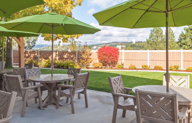 Outdoor patio area with round wooden tables and chairs under large green umbrellas, surrounded by a fenced garden with green grass, shrubs, and trees under a partly cloudy sky.