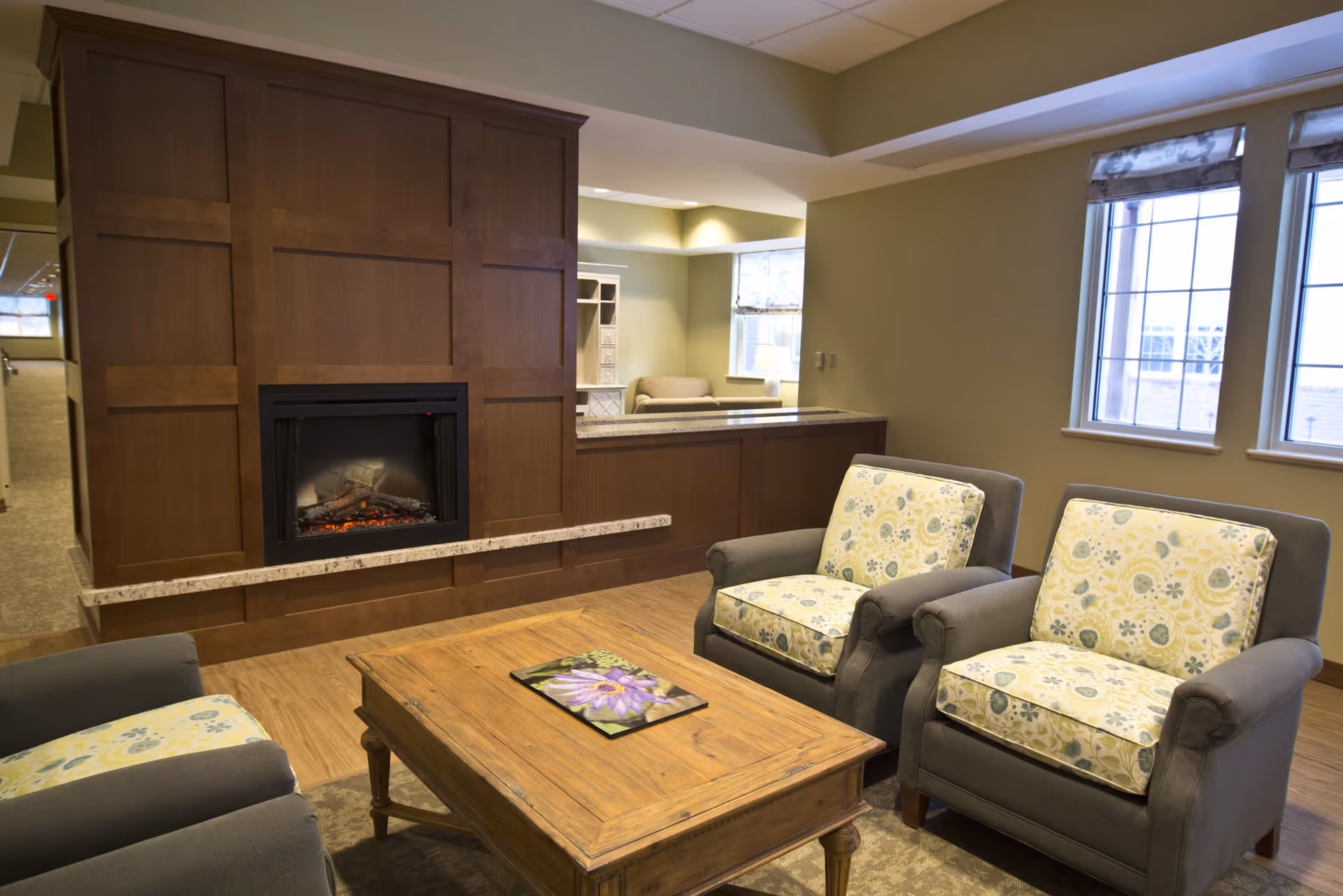 A cozy living room area with two gray armchairs featuring floral patterned cushions, a wooden coffee table with a book on it, a built-in electric fireplace with a wooden panel surround, and windows with patterned roman shades letting in natural light.