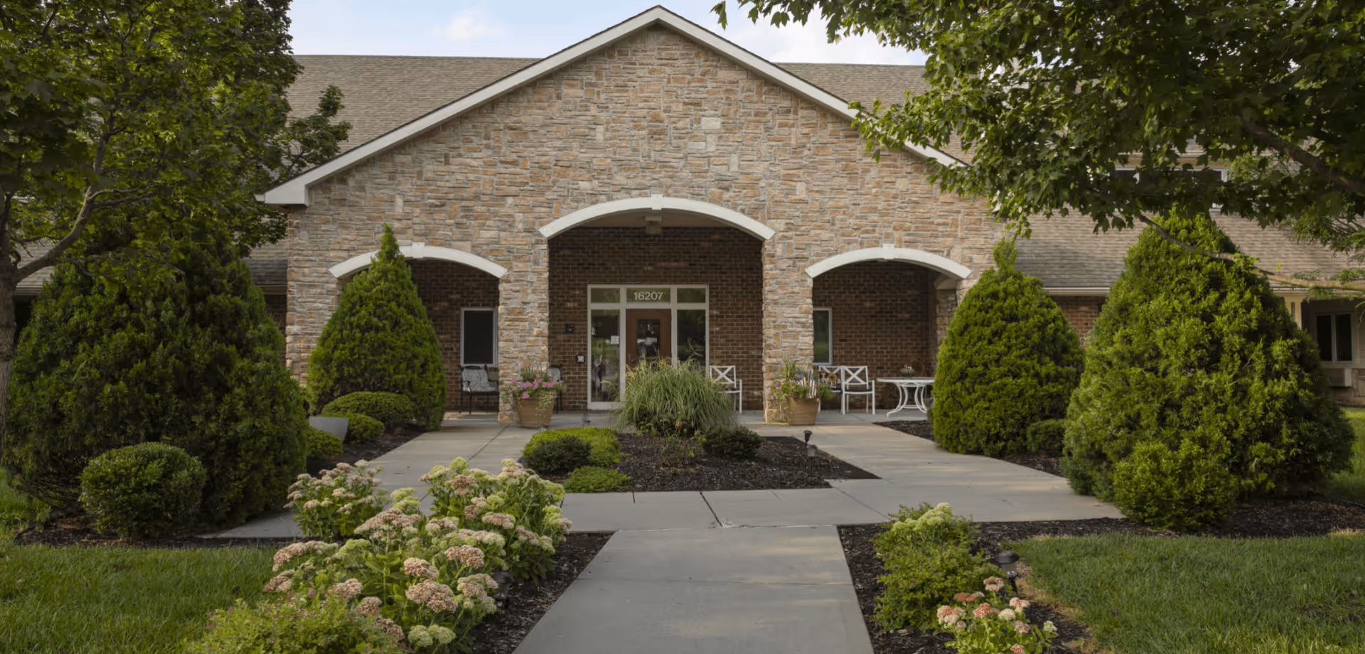 Front exterior view of a single-story assisted living facility with a stone and brick facade, a covered entrance with three arches, surrounded by neatly trimmed bushes, trees, and flower beds along a concrete walkway.