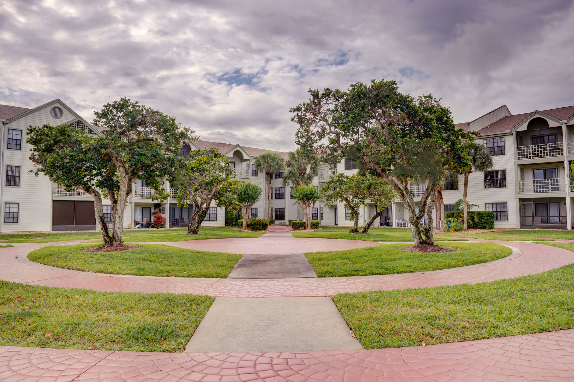 Courtyard with circular brick walkways, mature trees and a three-story senior living building in the background under a cloudy sky.